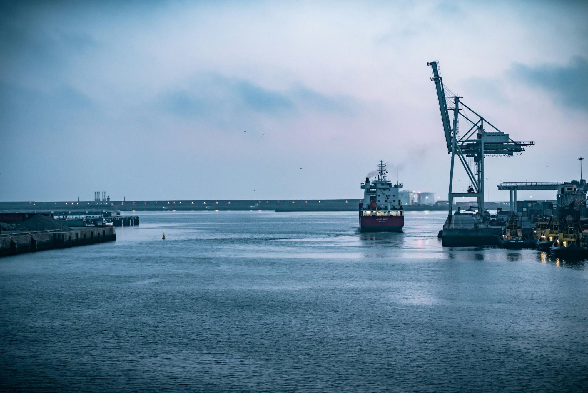A cargo vessel entering harbour at dusk past a quayside crane